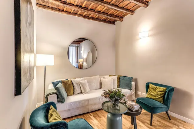 A staged living room with a beamed wooden ceiling, a cream sofa with cushions, two teal armchairs, a round wall mirror and a floor lamp.