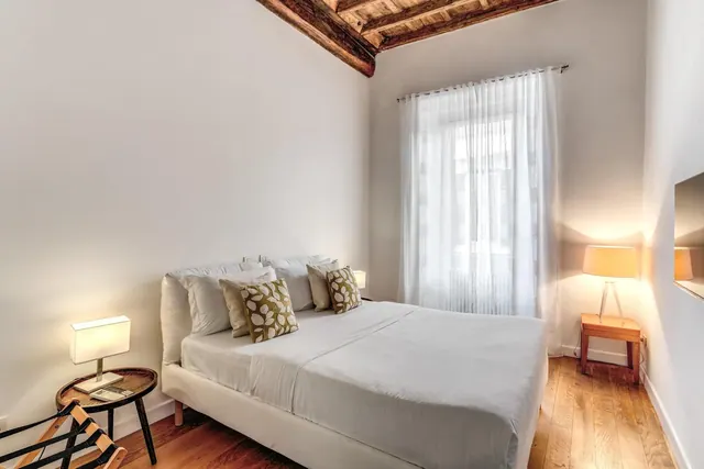 A double bed with white linen and patterned cushions sits beside a window with sheer curtains, with a lamp on a side table and exposed wooden beams on the ceiling.