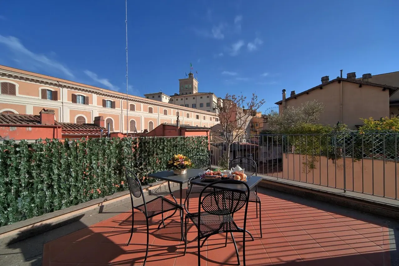 A rooftop terrace with a red tiled floor, black metal table and chairs set with tea and food, a faux-ivy screen, and a view of surrounding buildings under a clear blue sky.