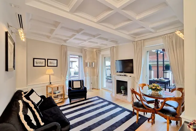 A living room with a coffered ceiling, black sofa and armchair, striped rug, fireplace, wall-mounted television and a dining table by open doors to a balcony.