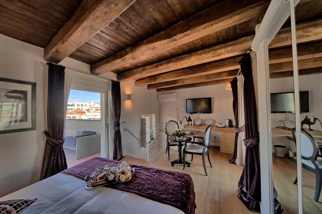 A bedroom with a wooden beamed ceiling, a bed with a purple throw and breakfast tray, a small sitting area and desk, and an open balcony door showing rooftops and buildings.