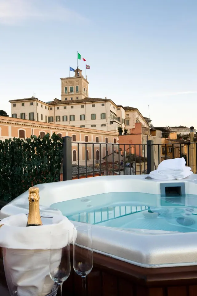 A rooftop terrace hot tub holds a champagne bottle in an ice bucket, two glasses and folded towels, with a view over city buildings and a large cream-coloured building topped with flags.