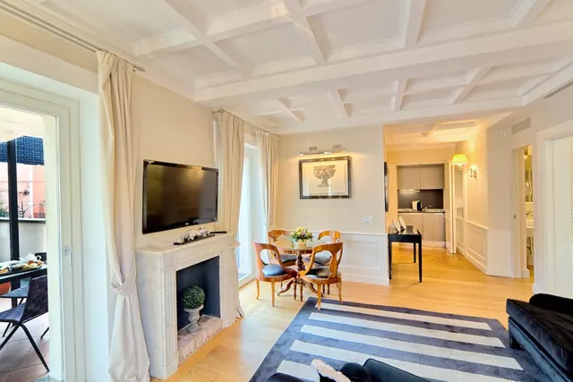A bright living room with a wall-mounted television above a marble fireplace, a small dining table with four chairs, a striped rug, and a kitchenette visible through an open doorway.