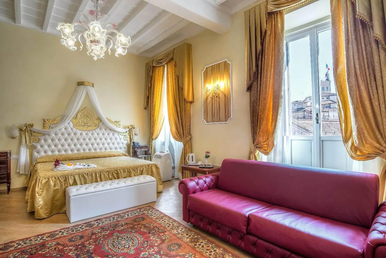 A bedroom with a gold tufted king bed, a red leather sofa, and a window overlooking Rome rooftops and a bell tower.