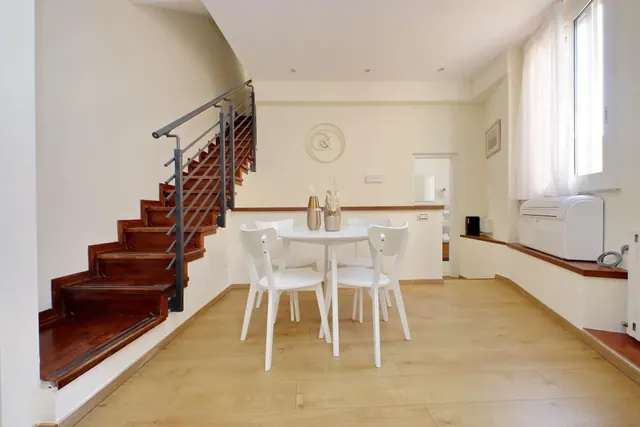 A bright dining area with a round white table and four white chairs, a wooden staircase with a metal railing on the left, and a wall-mounted air conditioning unit beneath a window.
