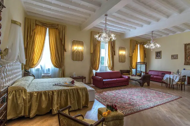 A large suite bedroom with a double bed, gold curtains, red leather sofas, crystal chandeliers and a dining table by the tall windows.