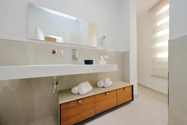 A bathroom with a long white wall-mounted vanity, a large framed mirror, a wall-mounted hair dryer, and rolled white towels on a wooden drawer unit.