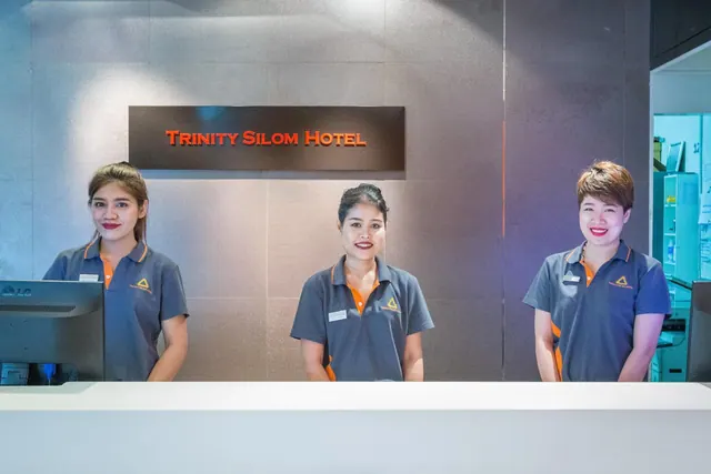 Three staff in grey polo shirts stand behind a white reception counter beneath a sign reading Trinity Silom Hotel.
