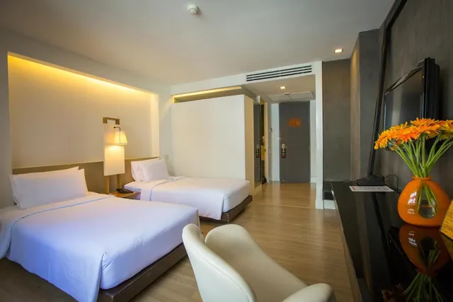 A hotel room with two white beds, a bedside lamp, a black desk holding a flat-screen TV and an orange vase of flowers, and two cream chairs in front of the desk.