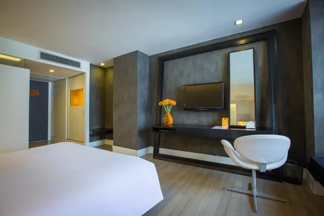 A bedroom with a large white bed, a wall-mounted television, a desk with a white swivel chair, and a vase of orange flowers.