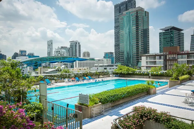 An outdoor rooftop lap pool with lane ropes and sun loungers, with tall glass towers rising behind it under a partly cloudy sky.