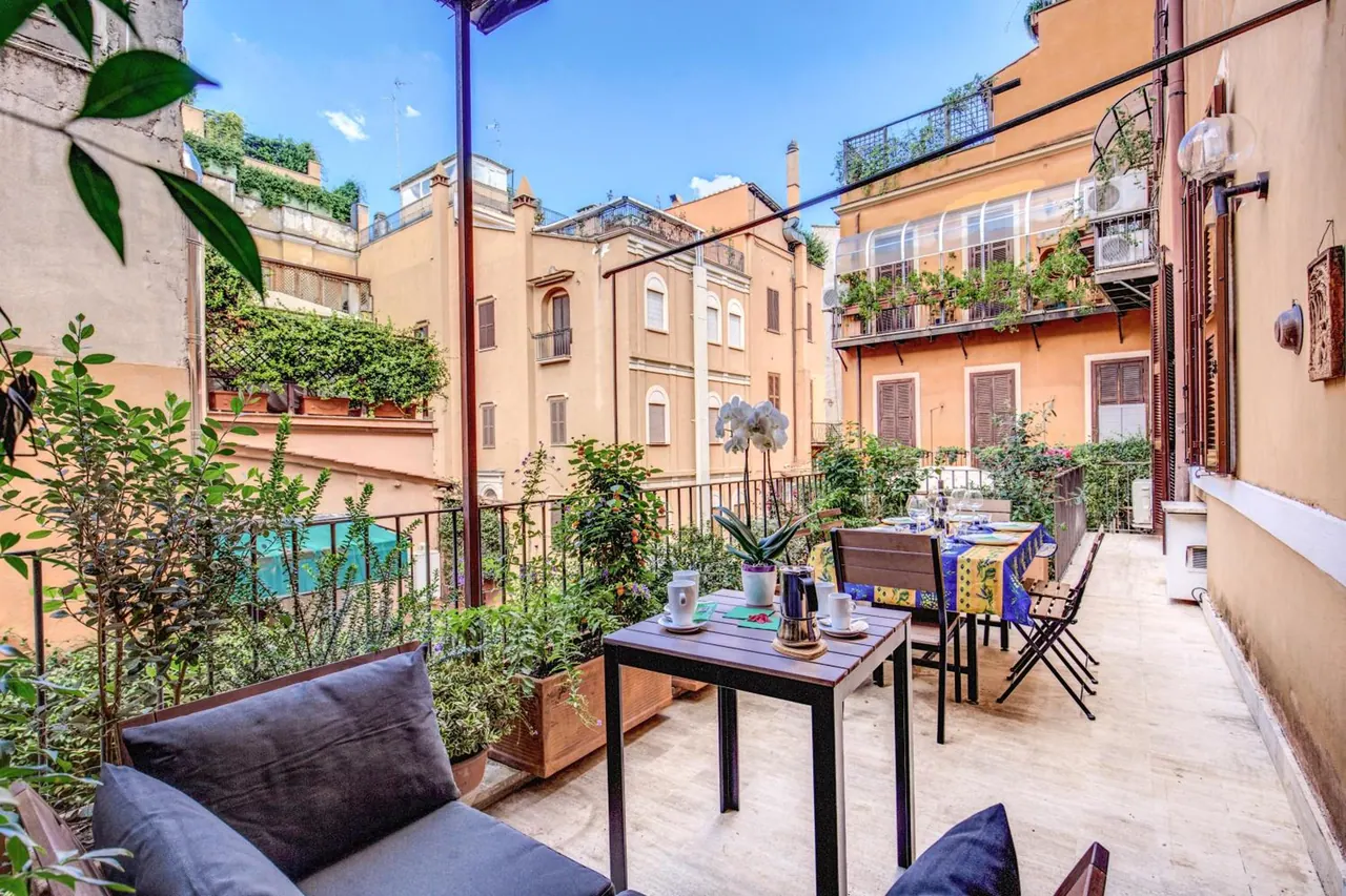 A plant-filled terrace with an outdoor sofa, a small table with coffee cups, and a dining table with a blue tablecloth, overlooking neighbouring buildings.