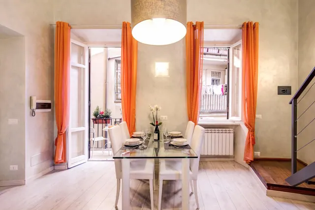 A dining area with a glass table set for two, white chairs, orange curtains, and an open door to a small balcony with a chair and plant planters.