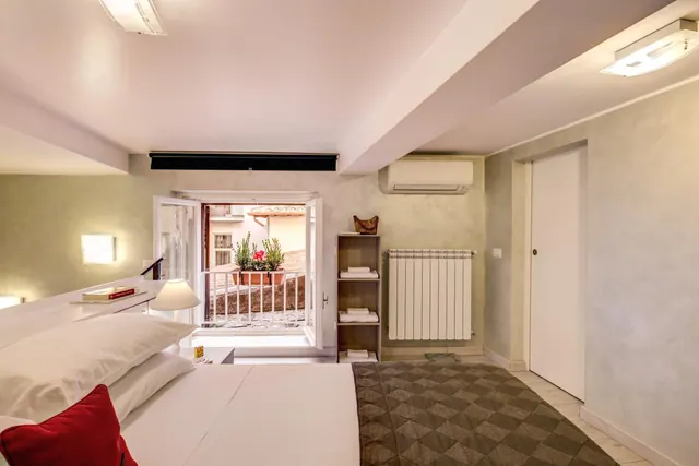 A bedroom with a made double bed, a radiator, a wall-mounted air conditioner, and French doors opening onto a small balcony with potted plants.