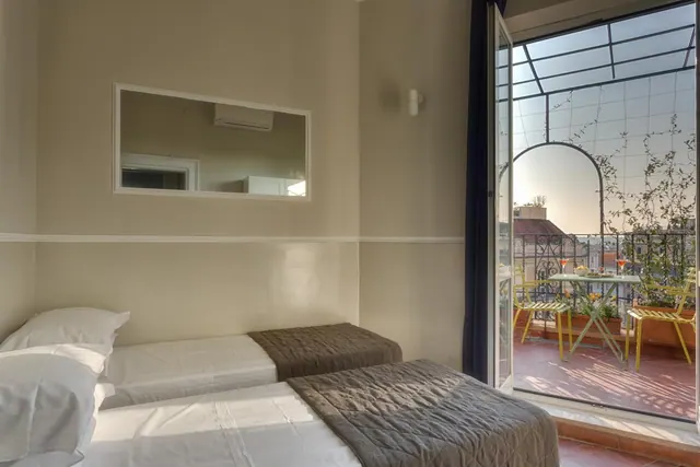 A bedroom with two single beds and a wall mirror, with an open door onto a terrace containing yellow chairs and a small table.
