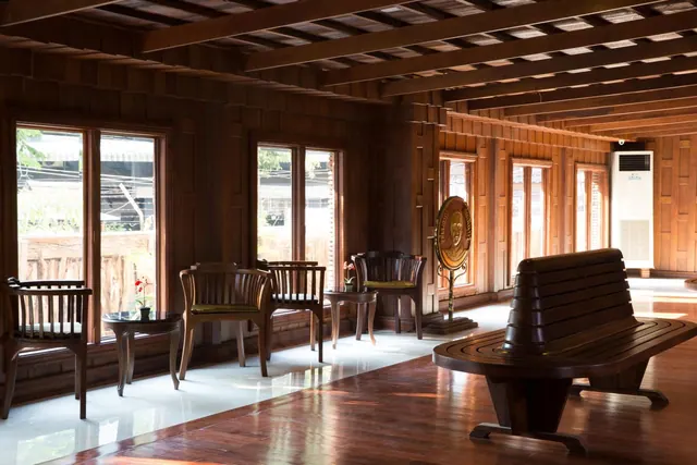 A wood-panelled seating area with teak armchairs, a long wooden bench, small side tables and a bronze disc on a stand, with windows and a wall-mounted air conditioner.