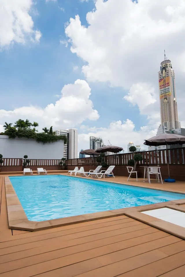 A rooftop swimming pool with a wooden deck, white sun loungers, umbrellas and a tall tower in the background under a cloudy sky.