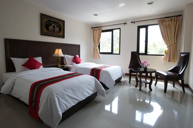 A hotel room with two twin beds made up with white bedding and red accents, two armchairs beside a small round table, and windows with curtains looking out onto trees.