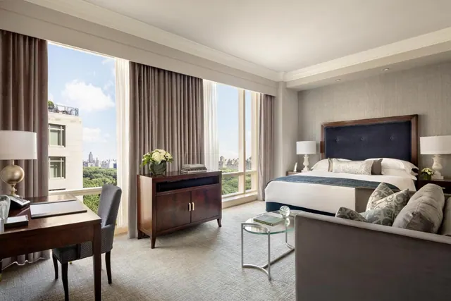 A hotel bedroom with a king bed, a desk, a sofa and large windows overlooking green trees and city buildings.