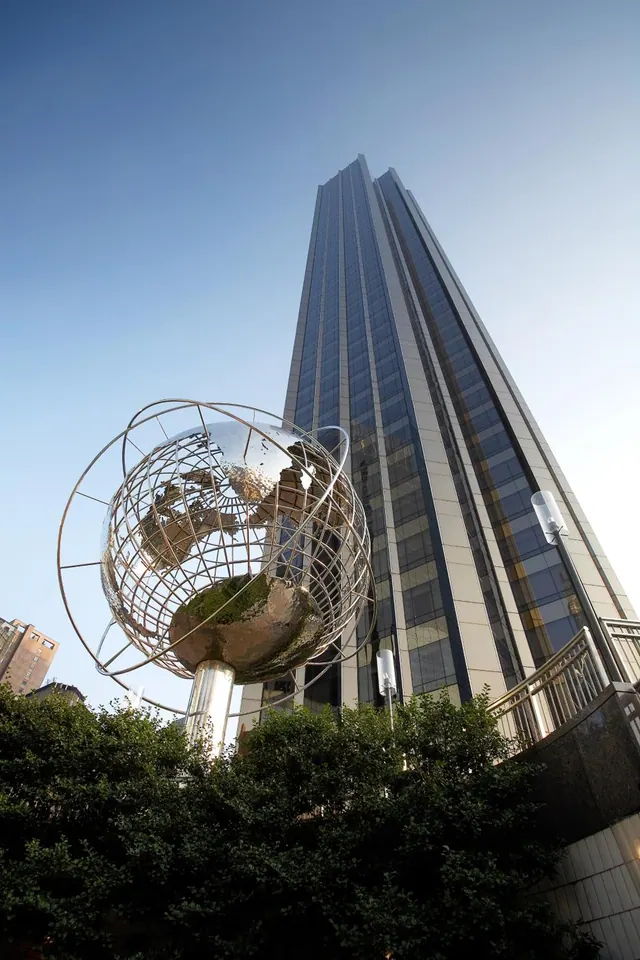 A tall glass-clad skyscraper rises behind a large metal armillary sphere sculpture and trees under a clear blue sky.
