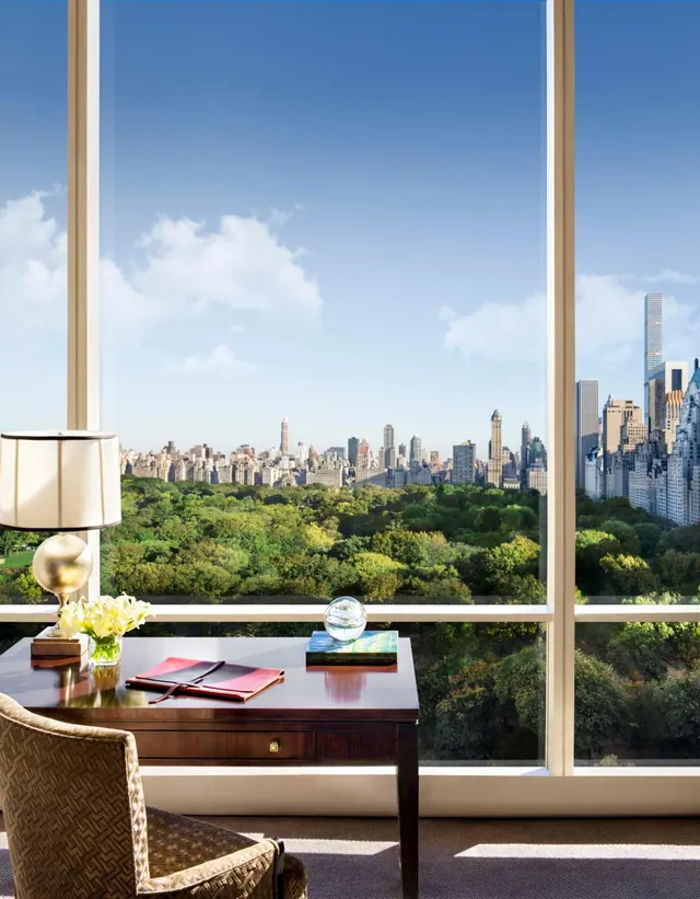 A wooden desk and upholstered armchair stand in front of floor-to-ceiling windows overlooking Central Park and the Manhattan skyline.