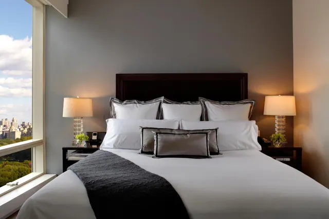 A king bed with white linens and grey cushions sits beside a large window overlooking city buildings and trees, with a lamp on each nightstand.