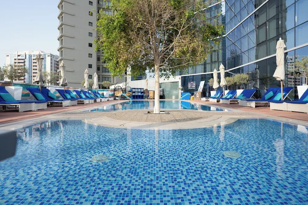 Outdoor pool with blue mosaic tiles and a tree growing in the water, with blue sun loungers and closed umbrellas on the deck and a glass-fronted building on the right.