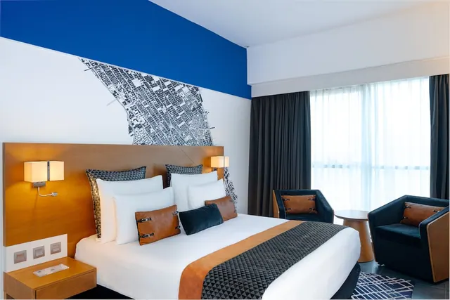 A bedroom with a made bed, a wall mural of a city map beneath a blue painted band, two armchairs and a tall window with sheer curtains.
