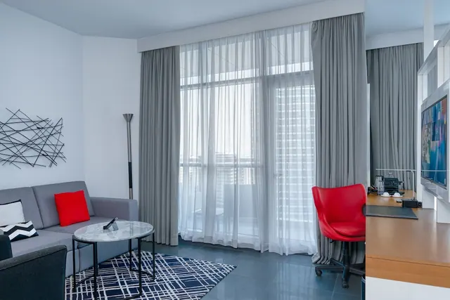 A living area with a grey sofa, a marble coffee table, a red leather desk chair and a wooden desk, with sheer-curtained floor-to-ceiling windows showing high-rise buildings outside.