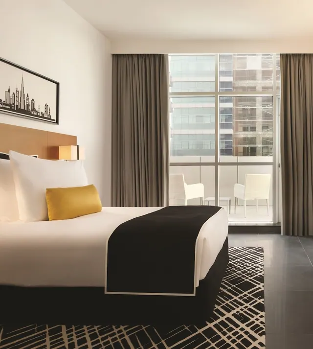 A bedroom with a made bed, a yellow accent cushion, a skyline artwork on the wall, and floor-to-ceiling glass doors opening onto a balcony with two white chairs.
