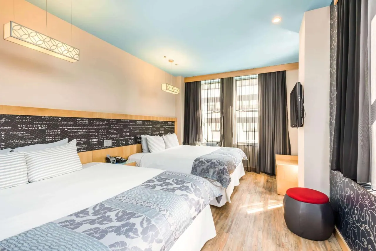 A hotel bedroom with two double beds, a wall-mounted television, a red ottoman and a pale blue painted ceiling.