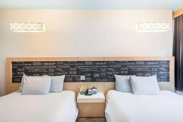 A hotel bedroom with two made beds, a headboard panel printed with New York text, pendant lights, and a bedside table holding a telephone and flowers.
