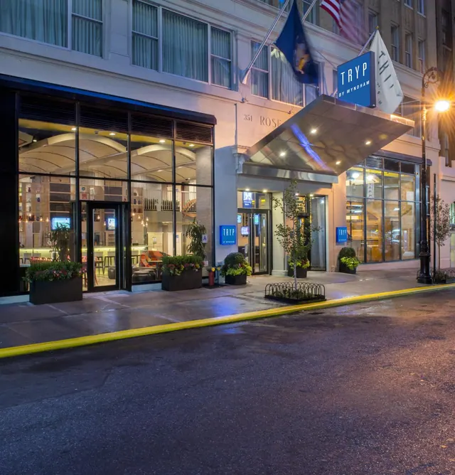 Exterior of the TRYP by Wyndham hotel entrance at dusk, with a metal canopy, potted plants and a street with a yellow curb in front.