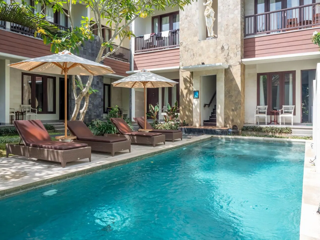 A rectangular swimming pool with sun loungers and parasol umbrellas sits in a courtyard beside a multi-storey building with balconies and a statue on the facade.