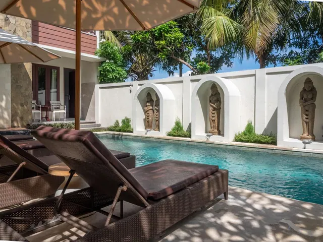 A long swimming pool with carved stone figures set in wall niches, with sun loungers and a parasol in the foreground.