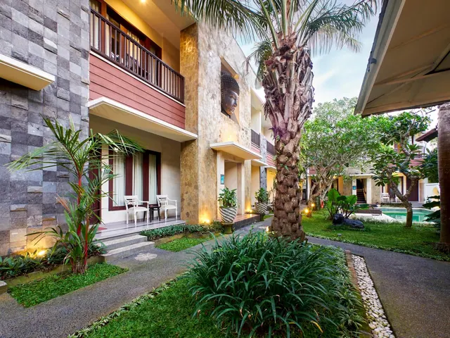 A two-storey courtyard building with an upper balcony, a Buddha head set in the stone wall, a palm tree, and a garden walkway with a small pool in the background.