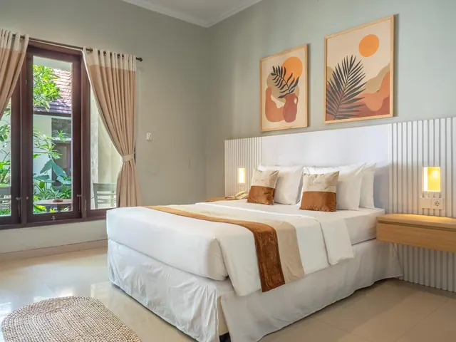 A large bed with white linen and a brown runner sits beside a window that looks out onto green foliage, with framed abstract art on the wall above.