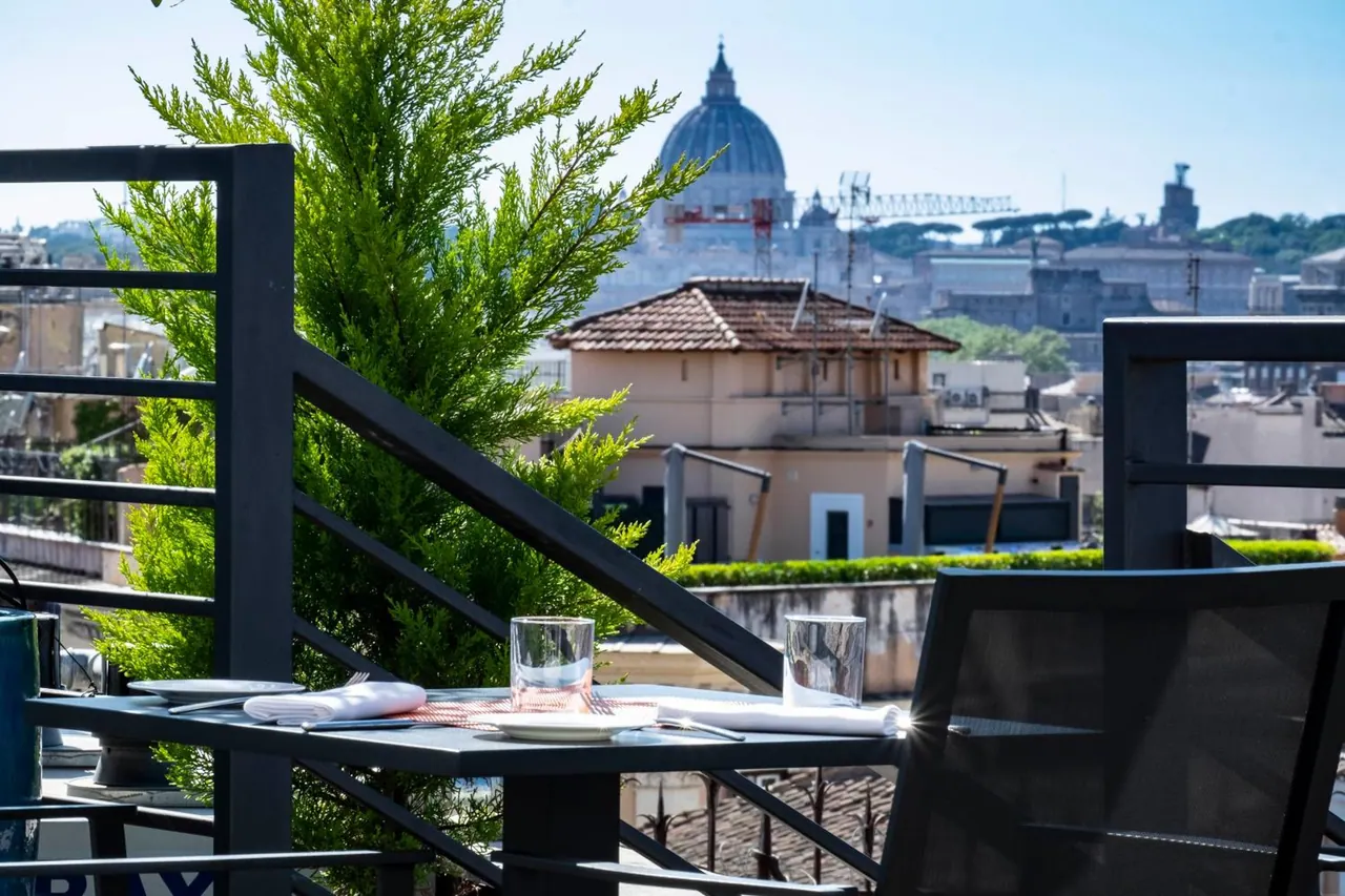A set outdoor table with napkins, glasses and a chair on a rooftop terrace, with the dome of St. Peter's Basilica and rooftops in the background.