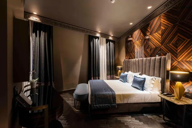 A bedroom with a large upholstered bed with blue cushions, dark curtains, a marquetry wood wall and a desk on the left.