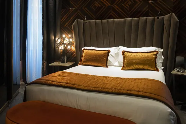 A double bed with a tall upholstered headboard, gold cushions and a brown throw, with a lit wall lamp and sheer curtains beside it.