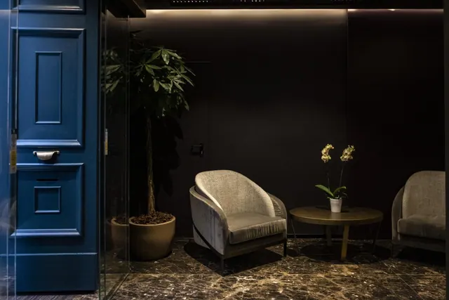 A lobby seating area with two upholstered armchairs, a round side table holding an orchid, a potted plant, and a blue panelled door beside a glass partition.