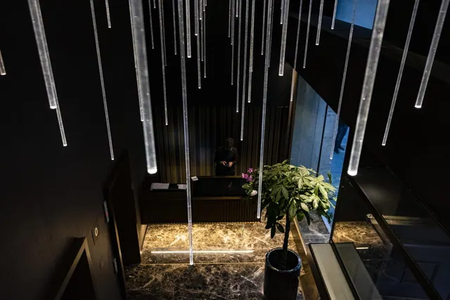 A dark lobby with suspended light tubes, a marble floor, a potted plant and a person seated at a reception desk.