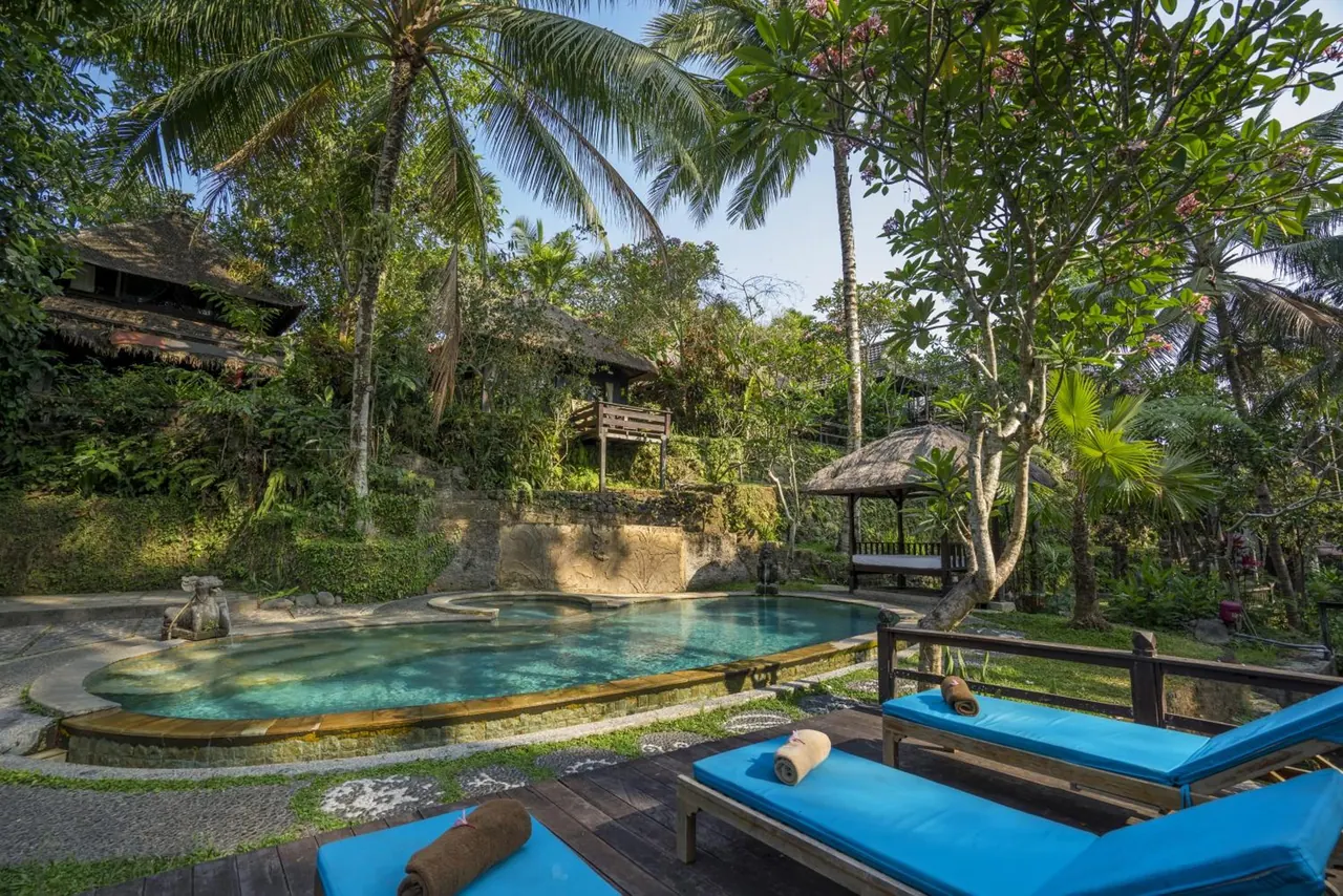 A swimming pool with blue sun loungers and rolled towels on a timber deck, surrounded by palm trees, tropical garden and a thatched gazebo.
