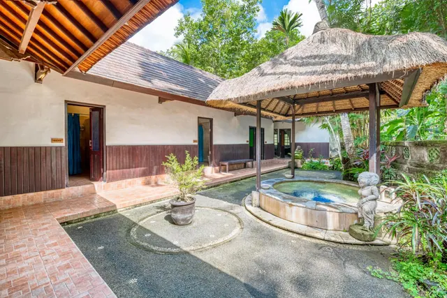 A small round plunge pool sits under a thatched gazebo beside a stone statue, with a villa building and a potted plant in the courtyard.