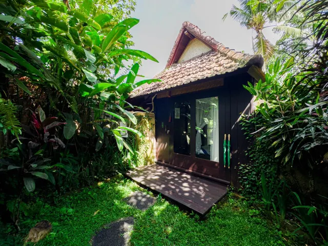 A small dark timber bungalow with a tiled roof and a wooden deck stands among tropical plants and grass.