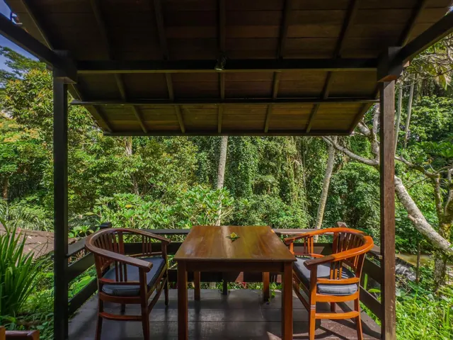 A covered wooden terrace holds a wooden table and two armchairs with dark cushions, overlooking dense green tropical trees.