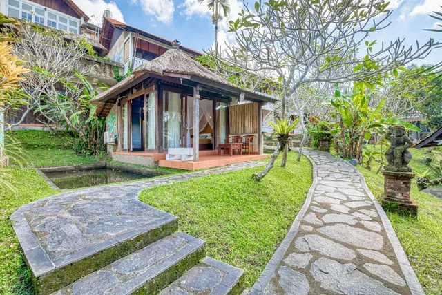 A single-storey thatched-roof bungalow with a small raised terrace stands beside a stone path through a garden with a pond and a stone statue.