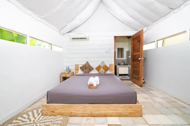 A bedroom with a wooden platform bed, batik cushions, folded white towels, a wall-mounted air conditioner, and an open wooden door to a bathroom with a stone sink.