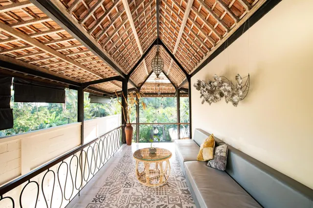 A covered balcony with a long grey sofa, a rattan coffee table and patterned floor tiles, with tropical greenery visible beyond the railing.