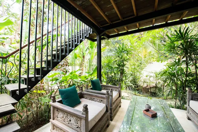 A covered terrace with two carved wooden armchairs with cream cushions and a glass-top table, open to a lush tropical garden, with a black metal staircase on the left.